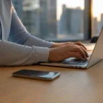 Händer som skriver på laptop med iPhone bredvid på skrivbord i kontorsmiljö